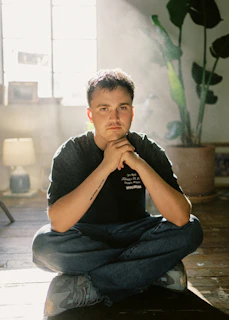 A calm, mindful individual wearing the stillpausez tee, sitting cross-legged in a sunlit room surrounded by plants.
