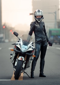 A person dressed in black leather motorcycle gear stands confidently beside a sleek sport bike on an empty urban street. The motorcycle features a distinctive design with bold graphics, and the rider is wearing a full-face helmet.