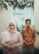 A woman in a pink hijab and dress, standing next to a man in a patterned shirt. They are posing in front of a decorative backdrop adorned with green foliage and colorful flowers.