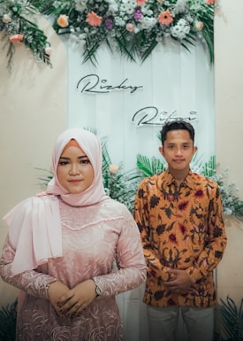 A woman in a pink hijab and dress, standing next to a man in a patterned shirt. They are posing in front of a decorative backdrop adorned with green foliage and colorful flowers.