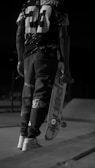 A person dressed in casual, urban attire holds a skateboard under one arm. The image is captured in black and white, emphasizing the high-contrast patterns on the clothing, including a prominent number '23' on the back of the shirt. The setting appears to be an outdoor skate park, indicated by the visible ramps and concrete surface.