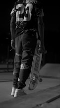 A person dressed in casual, urban attire holds a skateboard under one arm. The image is captured in black and white, emphasizing the high-contrast patterns on the clothing, including a prominent number '23' on the back of the shirt. The setting appears to be an outdoor skate park, indicated by the visible ramps and concrete surface.