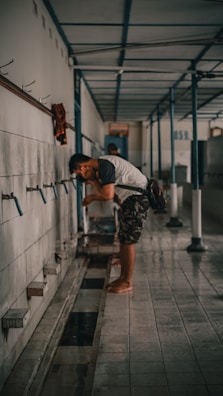 A man is bending over near a series of taps mounted on a tiled wall in a long corridor. The corridor has a tiled floor and a ceiling supported by metal beams. A wet area is visible on the floor, suggesting recent use of water. The man is wearing a white t-shirt and camo shorts. A bag hangs from his shoulder.