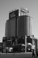 A large, cylindrical industrial building with the words 'Rec Room' on the front and a circular sign that is partially visible on top. Below, there is an old train engine and some industrial equipment. People are walking in the foreground, and a modern van is parked nearby. The scene is monochromatic.