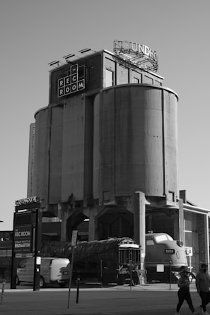A large, cylindrical industrial building with the words 'Rec Room' on the front and a circular sign that is partially visible on top. Below, there is an old train engine and some industrial equipment. People are walking in the foreground, and a modern van is parked nearby. The scene is monochromatic.