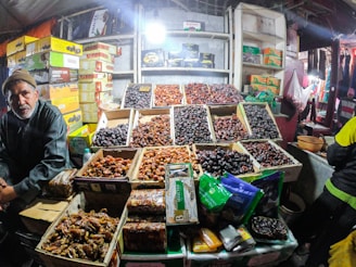 A market stall displaying a variety of dates neatly arranged in open boxes. A man, possibly the vendor, sits on the left side wearing a beanie and dark clothing. The background is populated with stacked boxes of different brands and packages of dates.