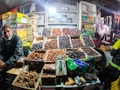 A market stall displaying a variety of dates neatly arranged in open boxes. A man, possibly the vendor, sits on the left side wearing a beanie and dark clothing. The background is populated with stacked boxes of different brands and packages of dates.
