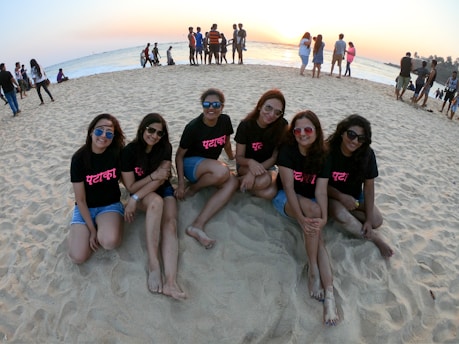 A group of six young women sitting together on a sandy beach, all wearing black t-shirts with bright pink text and sunglasses. Behind them, several people are walking along the shoreline, with the ocean and a setting sun in the background.