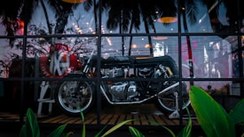 A motorcycle is displayed behind a large glass window with reflections of palm trees and interior lights. The setting appears to be a showroom or a stylish café, blending both natural and urban elements seamlessly.