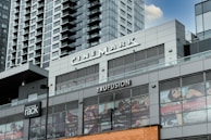 A modern commercial building featuring various business establishments, including a cinema and a fitness studio. The facade displays signage for Cinemark and Trufusion, with glass windows showing images related to fitness activities. Above the establishments are residential or office high-rise structures with numerous balconies.