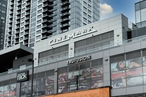 A modern commercial building featuring various business establishments, including a cinema and a fitness studio. The facade displays signage for Cinemark and Trufusion, with glass windows showing images related to fitness activities. Above the establishments are residential or office high-rise structures with numerous balconies.