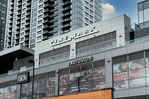 A modern commercial building featuring various business establishments, including a cinema and a fitness studio. The facade displays signage for Cinemark and Trufusion, with glass windows showing images related to fitness activities. Above the establishments are residential or office high-rise structures with numerous balconies.