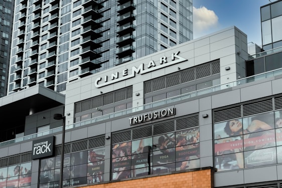 A modern commercial building featuring various business establishments, including a cinema and a fitness studio. The facade displays signage for Cinemark and Trufusion, with glass windows showing images related to fitness activities. Above the establishments are residential or office high-rise structures with numerous balconies.