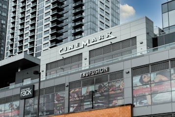 A modern commercial building featuring various business establishments, including a cinema and a fitness studio. The facade displays signage for Cinemark and Trufusion, with glass windows showing images related to fitness activities. Above the establishments are residential or office high-rise structures with numerous balconies.