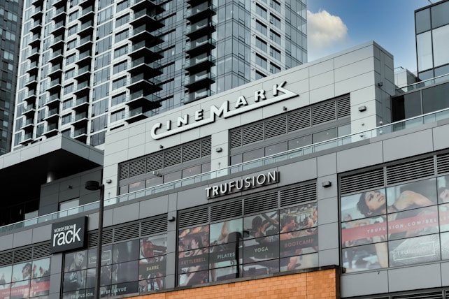 A modern commercial building featuring various business establishments, including a cinema and a fitness studio. The facade displays signage for Cinemark and Trufusion, with glass windows showing images related to fitness activities. Above the establishments are residential or office high-rise structures with numerous balconies.