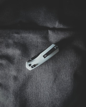 A folding pocket knife with a textured white handle and a black clip rests on a dark fabric surface. The lighting is dramatic, casting shadows and highlighting the texture of both the knife and the fabric.