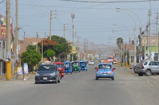 A busy urban street with various vehicles, including cars and three-wheeled tuk-tuks, driving along the road. The street is lined with utility poles, trees, and buildings, some of which display signs. The atmosphere appears sunny and clear, with mountains visible in the distant background.
