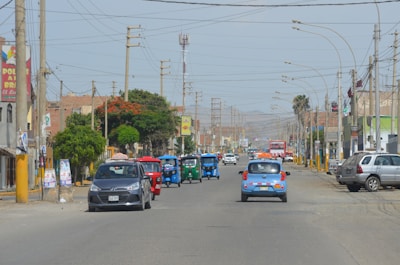 A busy urban street with various vehicles, including cars and three-wheeled tuk-tuks, driving along the road. The street is lined with utility poles, trees, and buildings, some of which display signs. The atmosphere appears sunny and clear, with mountains visible in the distant background.