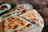 A wooden cutting board holds freshly made quesadillas, cut into slices, showcasing a filling of melted cheese, red bell peppers, and onions. An avocado is sliced open with one half visible, displaying its seed. A pizza cutter lies nearby on the board.
