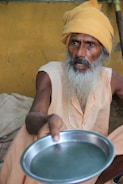 An elderly person with a long white beard is wearing a light orange robe and a matching turban. They are holding out a metal plate towards the viewer. The background is a plain yellow wall.