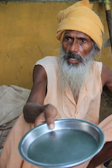 An elderly person with a long white beard is wearing a light orange robe and a matching turban. They are holding out a metal plate towards the viewer. The background is a plain yellow wall.