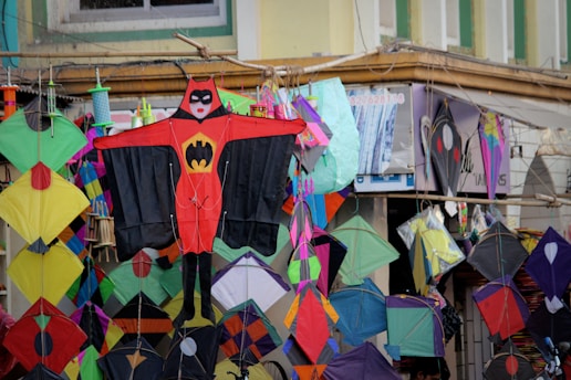 A vibrant display of various colorful kites is hanging in front of a shop. The kites feature different shapes and patterns, with one prominent kite in the center designed like a superhero costume. These kites are arranged closely, creating a lively and festive atmosphere.