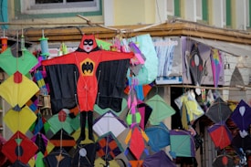 A vibrant display of various colorful kites is hanging in front of a shop. The kites feature different shapes and patterns, with one prominent kite in the center designed like a superhero costume. These kites are arranged closely, creating a lively and festive atmosphere.