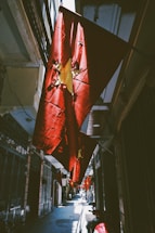 A narrow alleyway decorated with several red flags featuring a yellow star, indicative of the Vietnamese national flag. The flags are illuminated by sunlight, creating dramatic shadows and highlights on the walls and ground. The scene features a series of buildings on both sides, typical of an urban environment in Vietnam. Motorbikes are parked along the pathway.