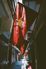 A narrow alleyway decorated with several red flags featuring a yellow star, indicative of the Vietnamese national flag. The flags are illuminated by sunlight, creating dramatic shadows and highlights on the walls and ground. The scene features a series of buildings on both sides, typical of an urban environment in Vietnam. Motorbikes are parked along the pathway.