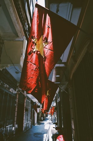 A narrow alleyway decorated with several red flags featuring a yellow star, indicative of the Vietnamese national flag. The flags are illuminated by sunlight, creating dramatic shadows and highlights on the walls and ground. The scene features a series of buildings on both sides, typical of an urban environment in Vietnam. Motorbikes are parked along the pathway.