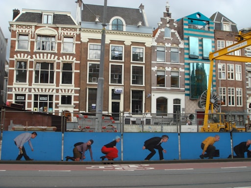 A row of traditional and modern buildings with a mural on a blue construction fence in front. The mural depicts various figures in a crouching position, resembling an evolution sequence. A yellow crane is visible on the right side, positioned next to a modern building with teal accents. The architecture features ornate gables and large windows.