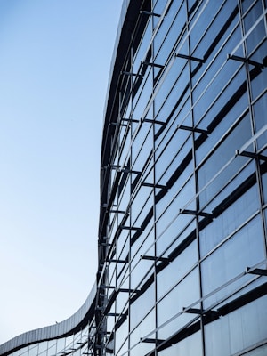A modern glass building with a curved facade featuring multiple reflective windows and metal framework. The architectural design emphasizes symmetry and sleekness, with a clear blue sky in the background.