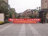 A large red banner with Chinese text is displayed across the entrance of a residential complex. The area is surrounded by modern brick apartment buildings, and there are some trees and shrubs around. The ground is paved with large tiles, and there are flowers blooming on the left side of the image.