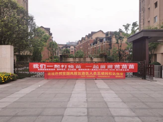 A large red banner with Chinese text is displayed across the entrance of a residential complex. The area is surrounded by modern brick apartment buildings, and there are some trees and shrubs around. The ground is paved with large tiles, and there are flowers blooming on the left side of the image.