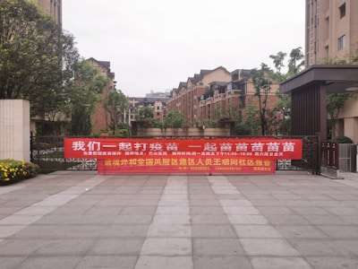 A large red banner with Chinese text is displayed across the entrance of a residential complex. The area is surrounded by modern brick apartment buildings, and there are some trees and shrubs around. The ground is paved with large tiles, and there are flowers blooming on the left side of the image.