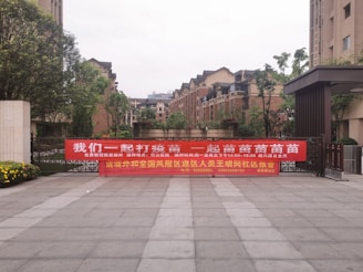 A large red banner with Chinese text is displayed across the entrance of a residential complex. The area is surrounded by modern brick apartment buildings, and there are some trees and shrubs around. The ground is paved with large tiles, and there are flowers blooming on the left side of the image.