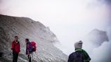 On a misty mountain slope, three hikers wearing outdoor gear and backpacks are engaged in a conversation. The background features a rugged, barren landscape partially obscured by fog.