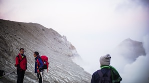 On a misty mountain slope, three hikers wearing outdoor gear and backpacks are engaged in a conversation. The background features a rugged, barren landscape partially obscured by fog.