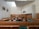 A large, formal courtroom featuring wooden furnishings, including benches, tables, and chairs arranged in a structured layout. The walls are light-colored with a high ceiling and ventilation panels. Artwork is displayed, including portraits on the walls and an emblem above the main area.