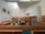 A large, formal courtroom featuring wooden furnishings, including benches, tables, and chairs arranged in a structured layout. The walls are light-colored with a high ceiling and ventilation panels. Artwork is displayed, including portraits on the walls and an emblem above the main area.