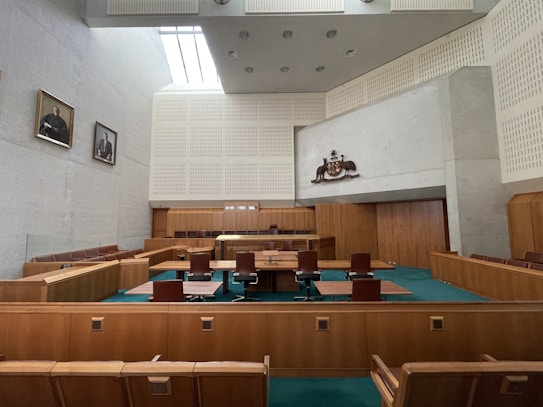 A large, formal courtroom featuring wooden furnishings, including benches, tables, and chairs arranged in a structured layout. The walls are light-colored with a high ceiling and ventilation panels. Artwork is displayed, including portraits on the walls and an emblem above the main area.