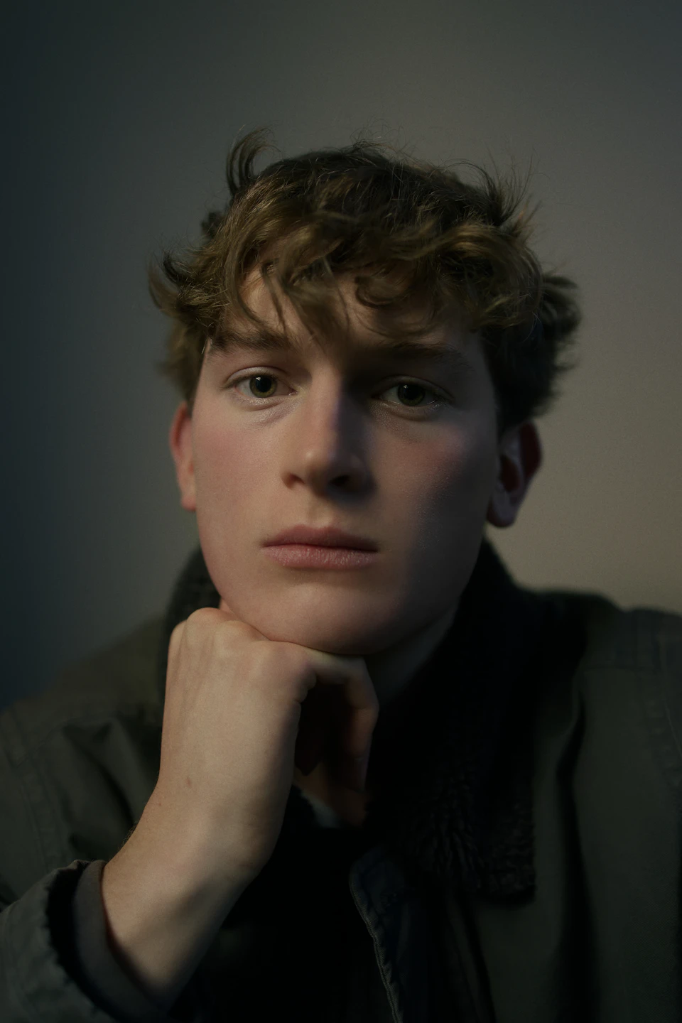 Young man with curly hair sits against a plain white wall, intensely staring forward