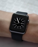 A black smartwatch with a square face is displayed on a person's wrist. The watch face shows a simple analog clock with white hour markers and hands, set against a dark background. The wrist is resting on a textured light-colored surface.