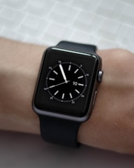 A black smartwatch with a square face is displayed on a person's wrist. The watch face shows a simple analog clock with white hour markers and hands, set against a dark background. The wrist is resting on a textured light-colored surface.
