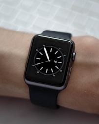 A black smartwatch with a square face is displayed on a person's wrist. The watch face shows a simple analog clock with white hour markers and hands, set against a dark background. The wrist is resting on a textured light-colored surface.