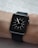 A black smartwatch with a square face is displayed on a person's wrist. The watch face shows a simple analog clock with white hour markers and hands, set against a dark background. The wrist is resting on a textured light-colored surface.