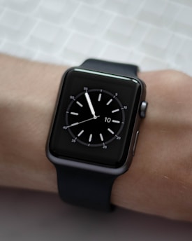 A black smartwatch with a square face is displayed on a person's wrist. The watch face shows a simple analog clock with white hour markers and hands, set against a dark background. The wrist is resting on a textured light-colored surface.
