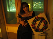 A person stands indoors holding a square painting featuring a circular design of red roses with green leaves and a banner across it bearing Cyrillic text. The setting appears to be at night, with dim lighting and city lights visible through a window.