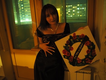 A person stands indoors holding a square painting featuring a circular design of red roses with green leaves and a banner across it bearing Cyrillic text. The setting appears to be at night, with dim lighting and city lights visible through a window.