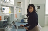 A person is standing in a laboratory, working on a laptop. The lab is equipped with various scientific instruments and glassware is visible on the shelves, including beakers and burettes containing different colored liquids. The atmosphere is well-lit with natural light coming from a large window.